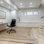 Bright modern basement office by basement contractors with white built-in cabinets, desk area, glass railing, light wood flooring, and recessed lighting.