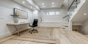 Bright modern basement office by basement contractors with white built-in cabinets, desk area, glass railing, light wood flooring, and recessed lighting.